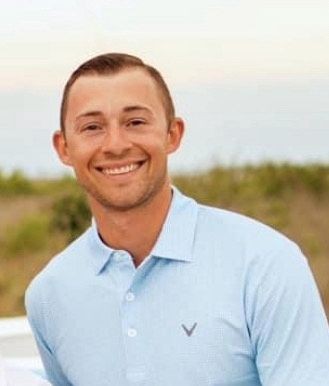 Man smiling and wearing a light blue collared shirt standing outdoors with a blurred natural background.