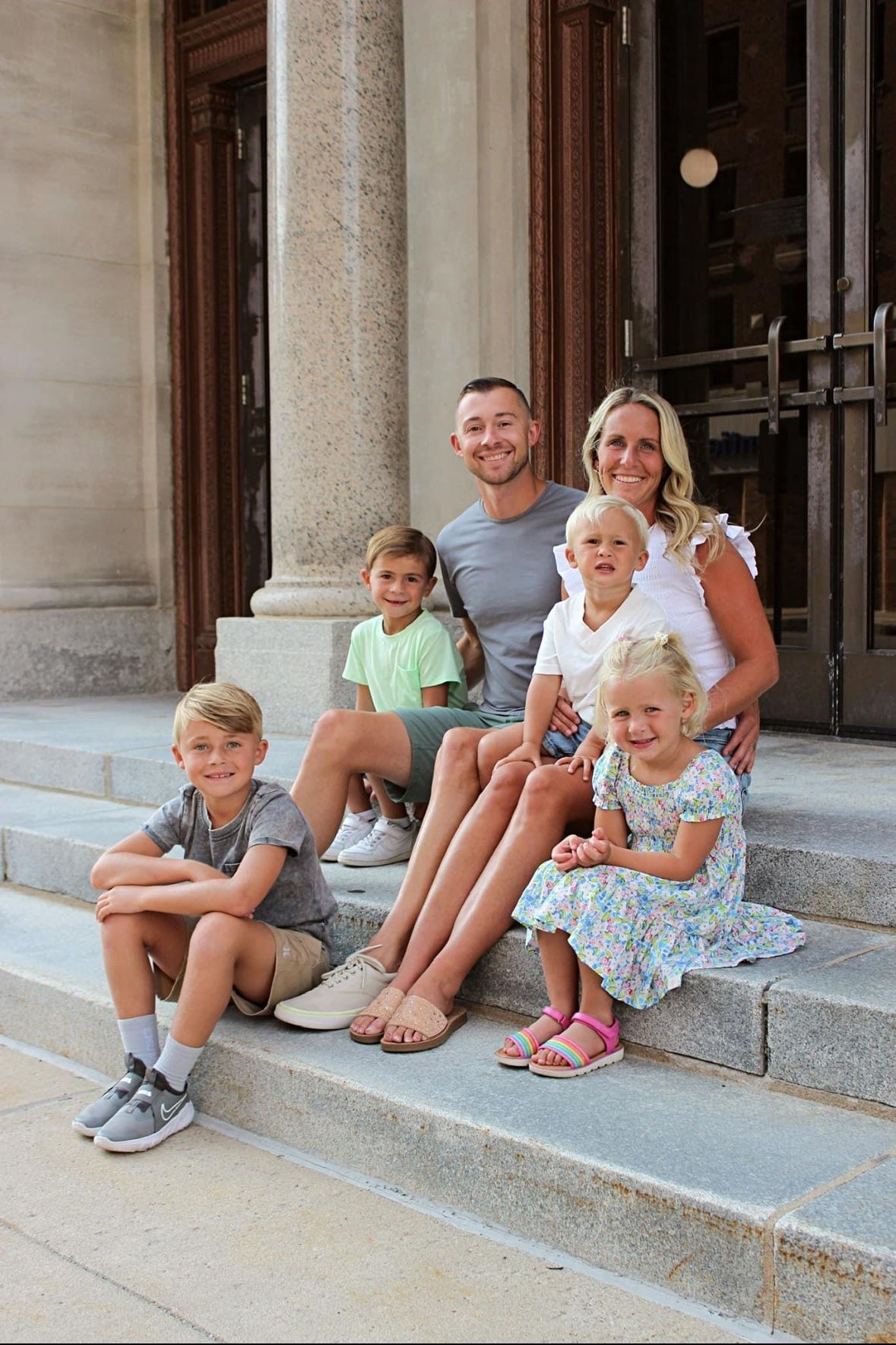 A family of six sitting on stone steps outside a building, smiling and posing for a picture.
